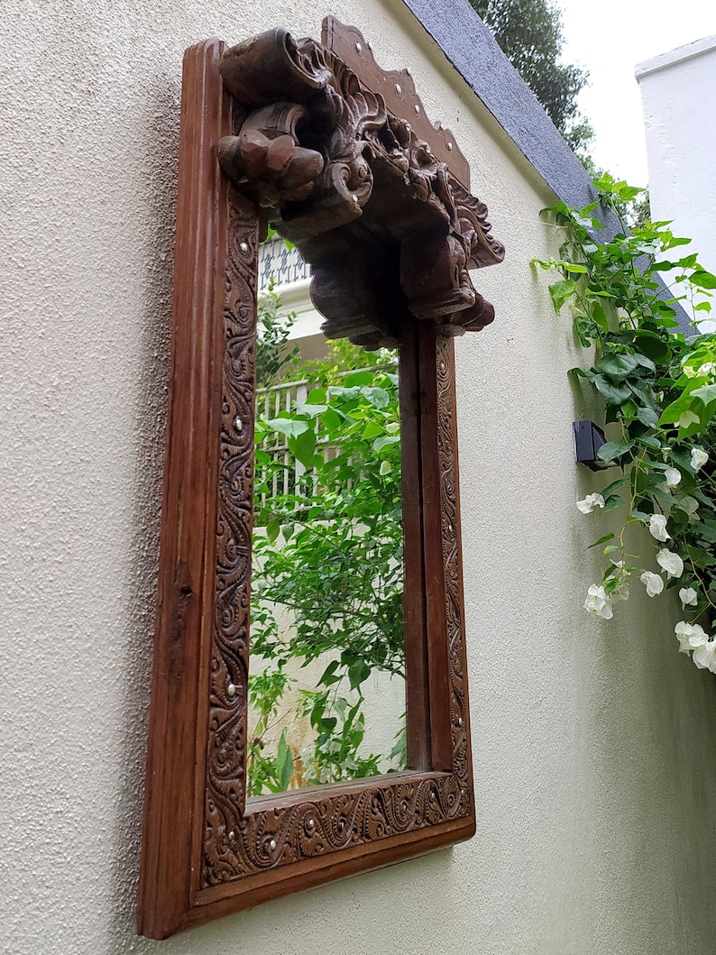 May include: A rectangular, ornate wooden mirror with a dark brown finish. The frame features intricate carvings and a decorative top. The mirror is mounted on a textured, off-white wall, reflecting greenery.