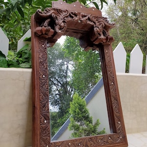 May include: A rectangular, ornate wooden mirror with intricate carvings and a decorative top. The frame is a rich brown color, with small, round embellishments. The mirror reflects a lush green outdoor scene.