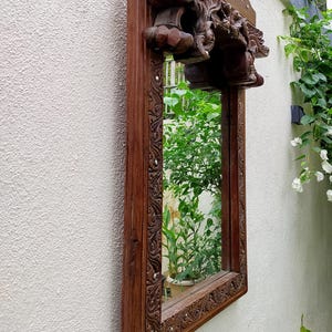 May include: A rectangular, wooden mirror with ornate carvings and a dark brown finish. The mirror is mounted on a textured, off-white wall, reflecting lush greenery. The top of the frame features detailed sculptural elements.