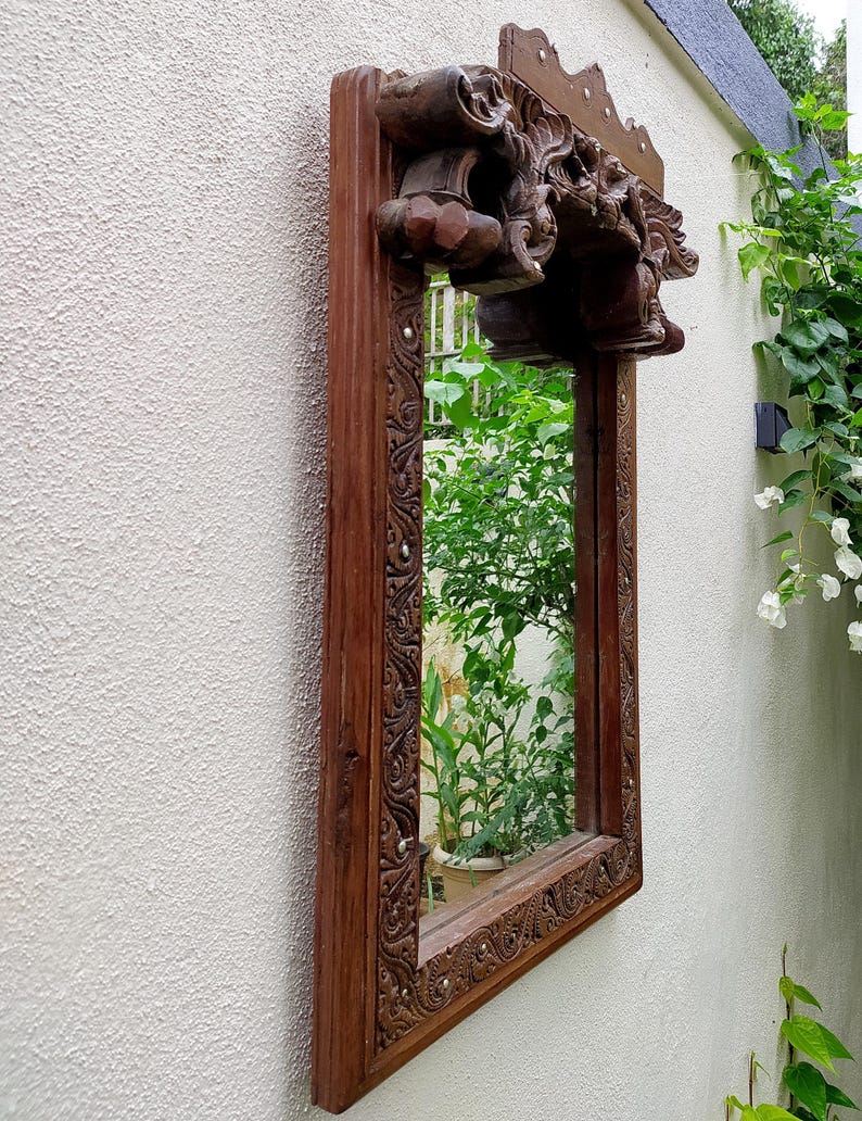 May include: A rectangular, wooden mirror with ornate carvings and a dark brown finish. The mirror is mounted on a textured, off-white wall, reflecting lush greenery. The top of the frame features detailed sculptural elements.