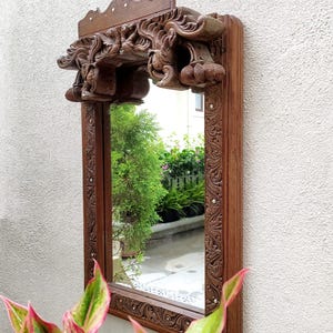 May include: A rectangular, wooden mirror with intricate carvings. The dark brown frame features detailed floral patterns and a decorative top with carved figures. The mirror is mounted on a white wall, with greenery visible in the reflection.