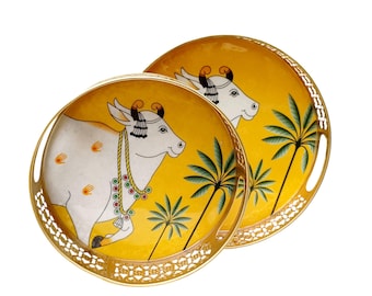 May include: Two round decorative trays featuring a yellow background and a white cow illustration. The trays have a gold filigree edge and handles. The cow is adorned with jewellery and palm trees are also depicted.