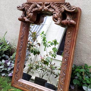 May include: A rectangular, ornate wooden mirror with a carved frame and decorative top. The mirror reflects plants in pots. The frame is a warm brown color, and the carvings add a vintage aesthetic. The mirror is leaning against a light-colored wall.
