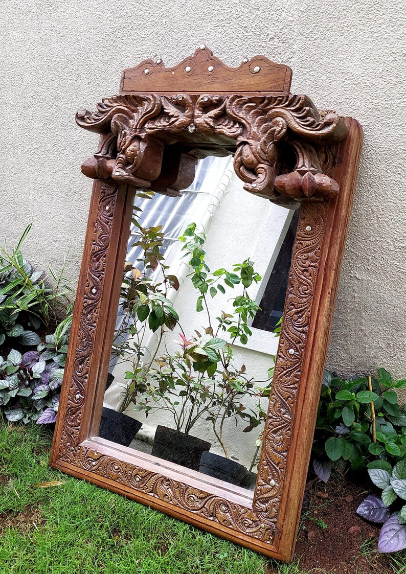 May include: A rectangular, ornate wooden mirror with a carved frame and decorative top. The mirror reflects plants in pots. The frame is a warm brown color, and the carvings add a vintage aesthetic. The mirror is leaning against a light-colored wall.