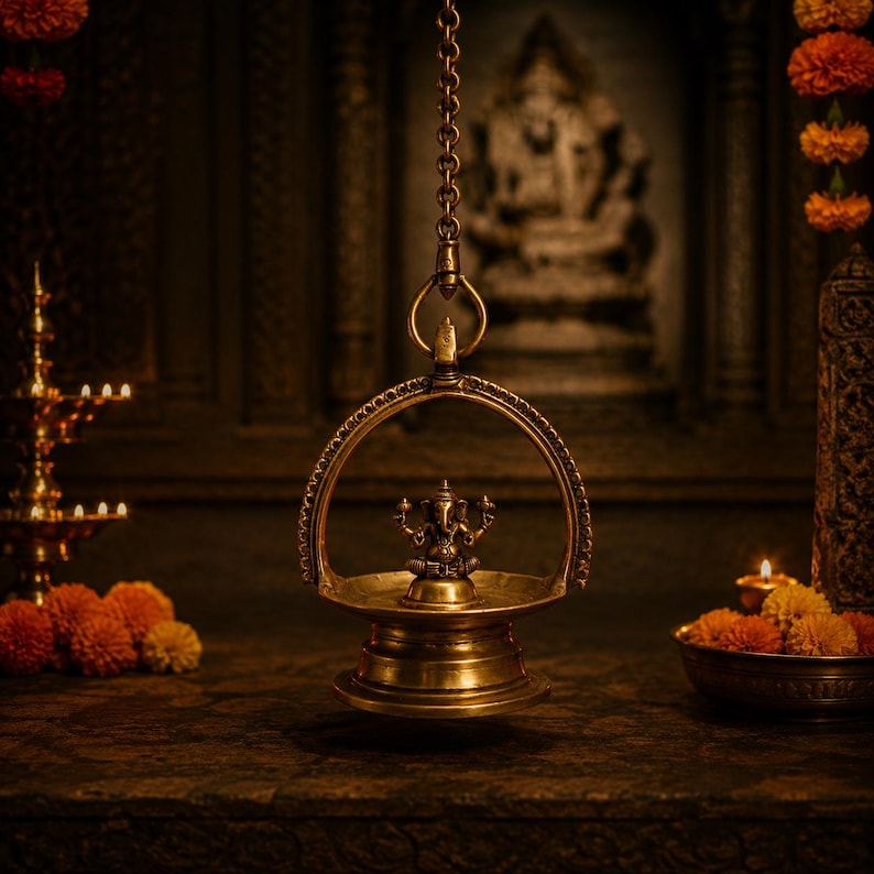 May include: A hanging brass oil lamp featuring a central Ganesha figurine within a circular frame, suspended by a metal chain. The lamp has a polished metallic finish and is positioned in a dimly lit space with warm ambient lighting and marigold flowers.