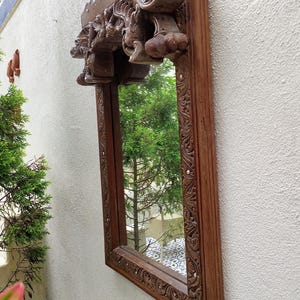 May include: A rectangular wooden mirror with intricate carvings and a decorative top. The frame is a warm brown color, and the mirror reflects a green tree. The carvings feature floral and abstract designs.