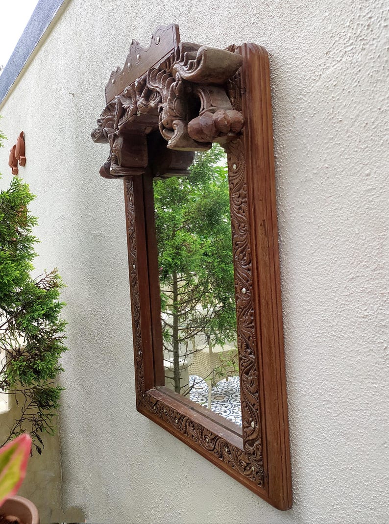 May include: A rectangular wooden mirror with intricate carvings and a decorative top. The frame is a warm brown color, and the mirror reflects a green tree. The carvings feature floral and abstract designs.
