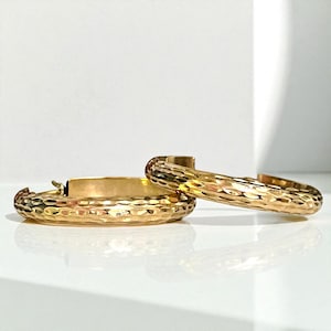 May include: A pair of gold-colored hoop earrings with a hammered texture. The earrings are displayed on a white surface, with one earring lying flat and the other angled upwards, showcasing the textured surface.