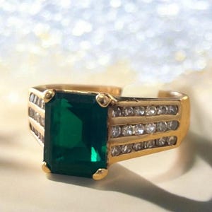 May include: Gold-toned ring featuring a large, emerald-cut green gemstone. The ring has a gold band with small, clear stones set along the sides. The ring is a statement piece, suitable for special occasions.