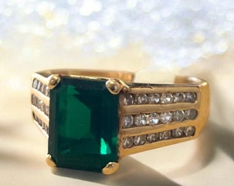 May include: Gold-toned ring featuring a large, emerald-cut green gemstone. The ring has a gold band with small, clear stones set along the sides. The ring is a statement piece, suitable for special occasions.