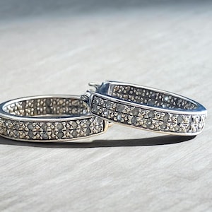 May include: A pair of silver-toned hoop earrings featuring a double row of small, clear, faceted stones embedded along the outer surface. The earrings have a hinged closure mechanism and a polished metallic finish that reflects light across the surface.