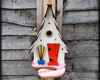 May include: A white birdhouse shaped like a house with a red door and a blue chair on a pink platform. The birdhouse has a black roof and a white chimney. The birdhouse is decorated with playing card symbols.