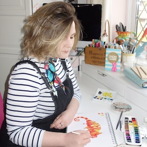 May include: A person wearing a striped shirt and overalls is drawing a red and orange chameleon on a white sketchbook. The sketchbook is open on a white desk with a watercolor palette and paintbrushes.