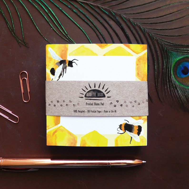 May include: A yellow and white memo pad with a honeycomb pattern and bee illustrations. The memo pad is made of 100% recycled paper and has 100 printed pages. The text on the memo pad reads "ALSO THE BISON Printed Memo Pad 100% Recycled - 100 Printed Pages - Made in the UK."