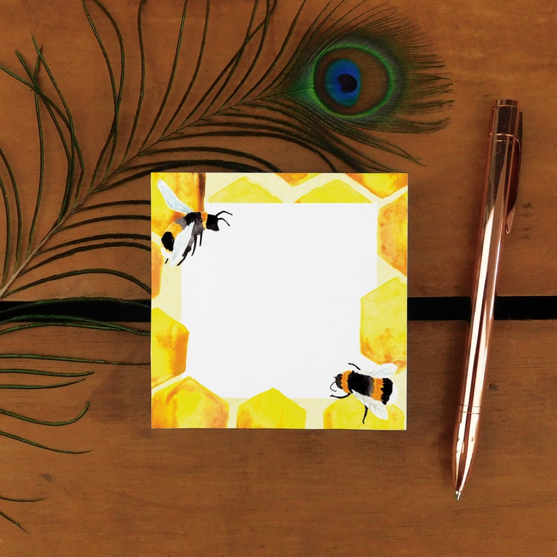 May include: A white notepad with a yellow honeycomb pattern and bee illustrations. The notepad is on a wooden surface with a peacock feather.