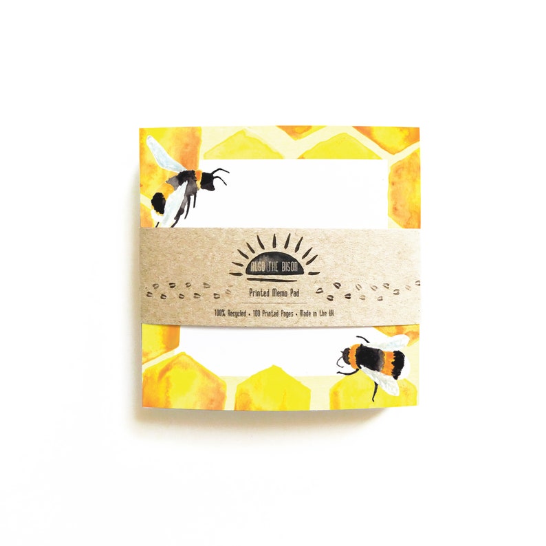 May include: A yellow and white memo pad with a watercolor design of honeycombs and bees. The memo pad is made of recycled paper and has 100 pages. The text on the memo pad reads "ALSO THE BISON Printed Memo Pad 100% Recycled - 100 Printed Pages - Made in the UK."