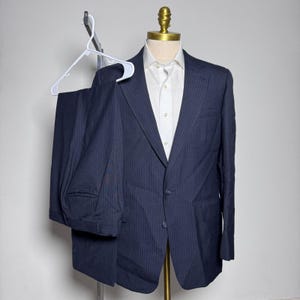 May include: A navy blue pinstripe suit displayed on a mannequin. The suit includes a two-button blazer and matching trousers, paired with a white dress shirt. The suit is hanging on a white hanger.
