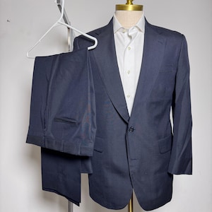 May include: A navy blue suit, including a blazer and trousers, displayed on a mannequin. The blazer has a single-button closure and is paired with a white dress shirt. The trousers are hanging on a white hanger.