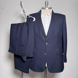 May include: A navy blue suit displayed on a mannequin and a hanger. The suit includes a two-button blazer with notched lapels and a pair of matching trousers. A white dress shirt is worn under the blazer. The suit is displayed against a plain white background.