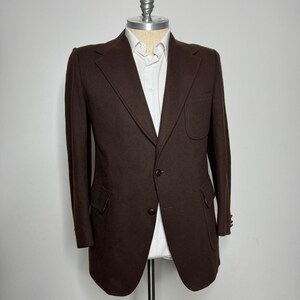 May include: A dark brown blazer with a two-button closure, notched lapels, and two front pockets. The blazer is displayed on a mannequin, revealing a white collared shirt underneath. The sleeves have three buttons at the cuff.