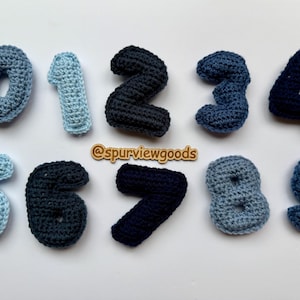 May include: A collection of ten crocheted numbers, ranging from zero to nine, in various shades of blue. The numbers are arranged on a white surface. The image includes the text "@spurviewgoods".