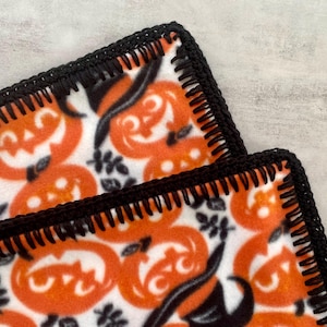May include: A white fleece blanket with a black crocheted border. The blanket features a repeating pattern of orange jack-o'-lantern faces.