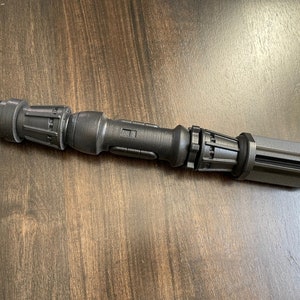 May include: A black metal lightsaber hilt with a textured surface and multiple sections. The hilt has a cylindrical shape and a rounded end.