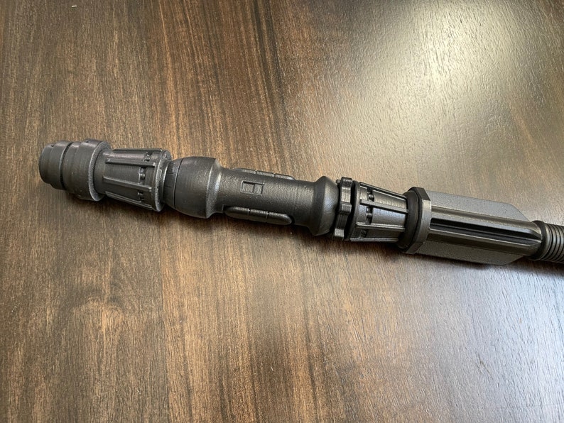 May include: A black metal lightsaber hilt with a textured surface and multiple sections. The hilt has a cylindrical shape and a rounded end.