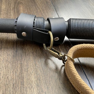 May include: Close-up of a black leather strap with a metal ring and a brown canvas strap attached to a black metal rod.