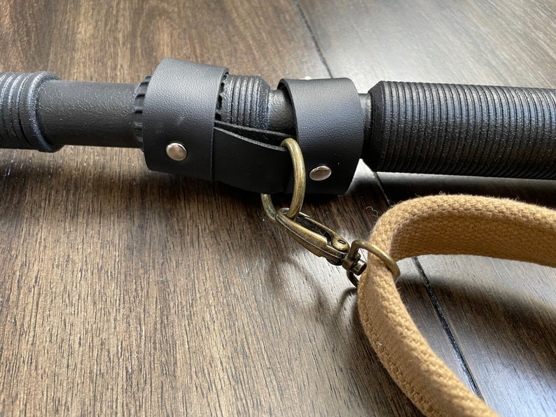 May include: Close-up of a black leather strap with a metal ring and a brown canvas strap attached to a black metal rod.