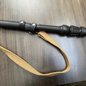 May include: A black telescopic baton with a brown canvas strap. The baton has a textured grip and a black plastic tip.