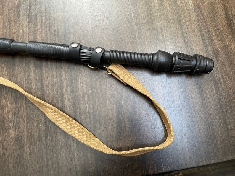 May include: A black telescopic baton with a brown canvas strap. The baton has a textured grip and a black plastic tip.