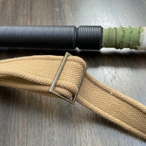 May include: A close-up of a brown canvas strap with a silver metal buckle. The strap is folded in half and the buckle is secured.