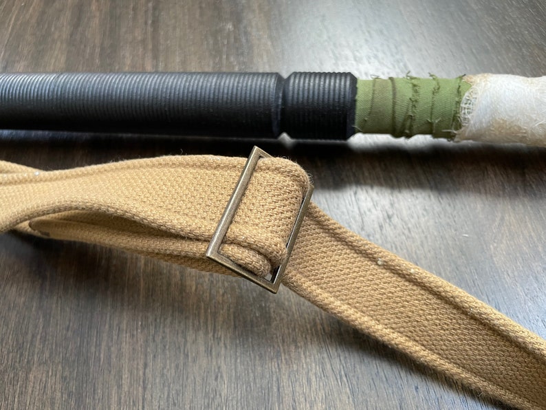 May include: A close-up of a brown canvas strap with a silver metal buckle. The strap is folded in half and the buckle is secured.