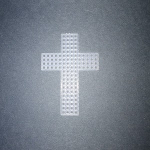 May include: A white plastic cross with a grid-like pattern. The cross is centered and stands out against a dark gray background. The design features a traditional Christian symbol.