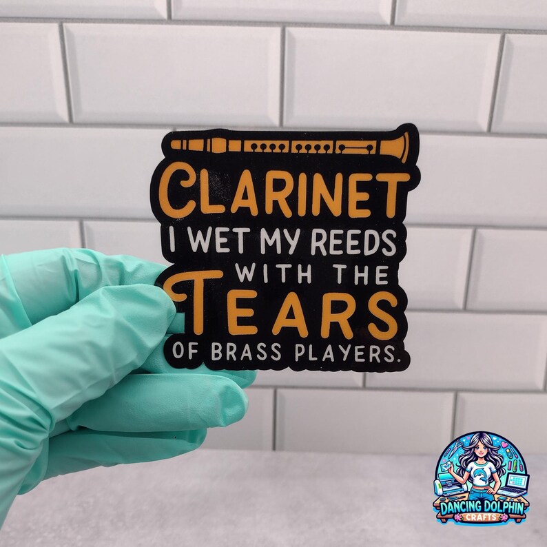 May include: A black sticker with a yellow graphic of a clarinet at the top. The text reads: CLARINET I WET MY REEDS WITH THE TEARS OF BRASS PLAYERS. The sticker has a die-cut shape and is held by a hand wearing a light blue nitrile glove.