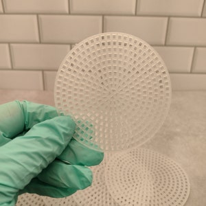 May include: A hand wearing a teal glove holds a white, circular plastic canvas sheet with a grid pattern. Additional sheets are visible in the background. The sheets are designed for crafting and needlepoint projects.