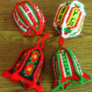 May include: Four handmade plastic canvas ornaments featuring festive red, green, white, and yellow yarn stitching. Two ornaments are bell-shaped with hanging loops, while the other two are rounded, spherical shapes with intricate geometric patterns.