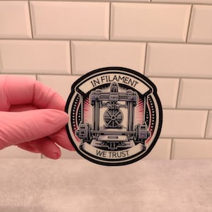 May include: A black and white sticker with a 3D printer design. The sticker says "In Filament We Trust" in a circular design.