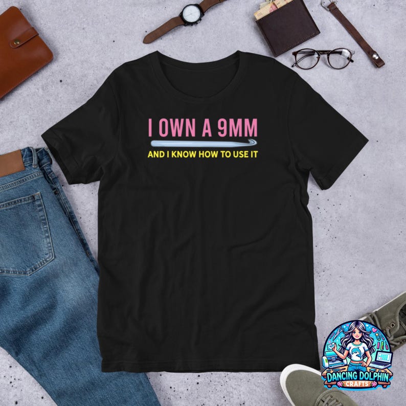 K&ouml;nnte beinhalten: Schwarzes T-Shirt mit dem Text "I own a 9mm and I know how to use it" in Pink und Gelb. Der Text befindet sich &uuml;ber einem d&uuml;nnen, hellblauen Rechteck.