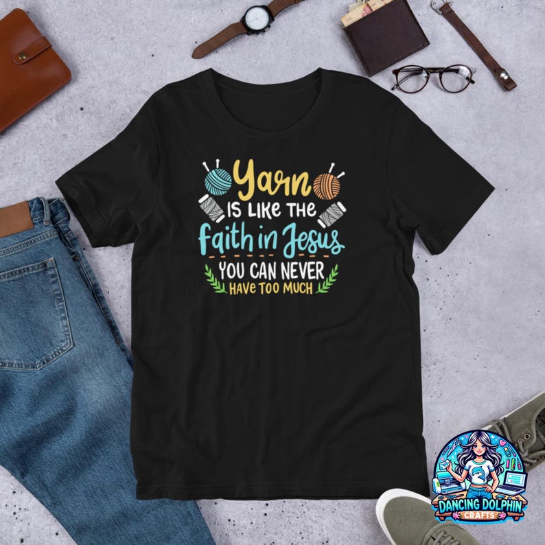 May include: Black t-shirt with the text "Yarn is like the faith in Jesus, you can never have too much." The design includes images of yarn and needles. The shirt is displayed on a flat surface with jeans and accessories.