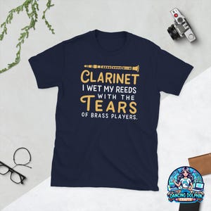 May include: A navy blue short-sleeve t-shirt featuring a graphic of a clarinet and the text: CLARINET I WET MY REEDS WITH THE TEARS OF BRASS PLAYERS. The design uses yellow and white lettering centered on the front of the garment.