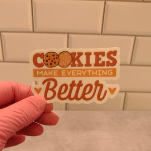 May include: White sticker with the text "COOKIES MAKE EVERYTHING BETTER" in orange and brown lettering. Two cookies are depicted in the text. The sticker has two small pink hearts on either side of the text.