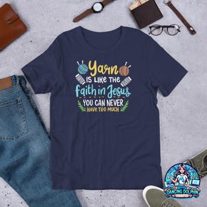 May include: Navy blue t-shirt with the text "Yarn is like the faith in Jesus, you can never have too much." The shirt features illustrations of yarn balls and needles. A logo for Dancing Dolphin Crafts is visible.