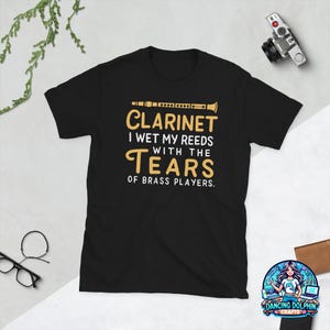 May include: A black short-sleeve t-shirt featuring a graphic of a clarinet and the text: CLARINET I WET MY REEDS WITH THE TEARS OF BRASS PLAYERS. The design is printed in gold and white ink on the front center of the garment.