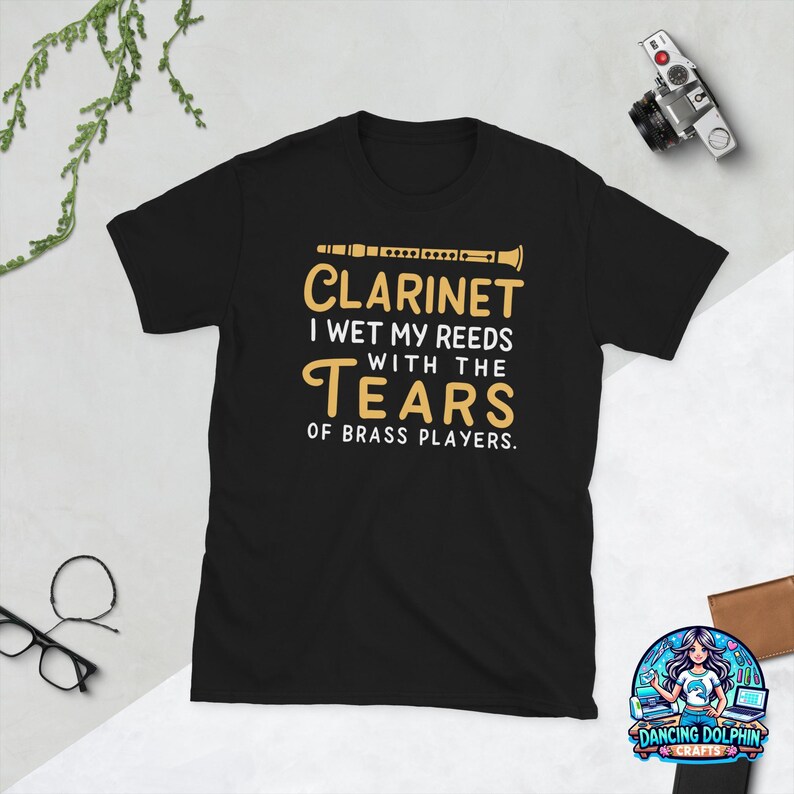 May include: A black short-sleeve t-shirt featuring a graphic of a clarinet and the text: CLARINET I WET MY REEDS WITH THE TEARS OF BRASS PLAYERS. The design is printed in gold and white ink on the front center of the garment.