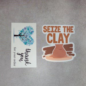 K&ouml;nnte beinhalten: Eine wei&szlig;e Karte mit einem blauen Schneeflockenbaumdesign und dem Text "Vielen Dank f&uuml;r Ihre Bestellung". Ein wei&szlig;er Aufkleber mit braunem Text, der "Seize the Clay" sagt, und ein brauner Tontopf auf einem wei&szlig;en Sockel.