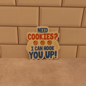 May include: A white sticker with blue, orange, and brown text and images. The text reads "Need Cookies? I can hook you up!" with three brown cookies pictured.