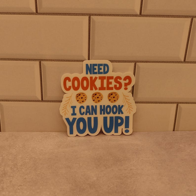 May include: A white sticker with blue, orange, and brown text and images. The text reads "Need Cookies? I can hook you up!" with three brown cookies pictured.