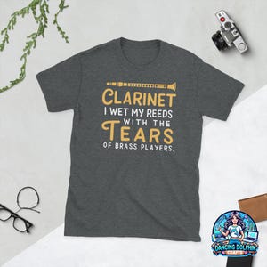 May include: A dark heather gray short-sleeve t-shirt featuring a graphic of a clarinet and the text: CLARINET I WET MY REEDS WITH THE TEARS OF BRASS PLAYERS. The design uses yellow and white lettering centered on the chest of the garment.